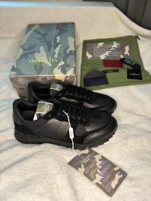 Svarta Valentino Garavani sneakers - Snygga svarta sneakers från Valentino Garavani med kamouflagemönster och detaljer i mocka och läder. Skorna har svarta skosnören, robust sula och coola nitar på hälen. Perfekta för dig som vill ha en exklusiv och trendig look.