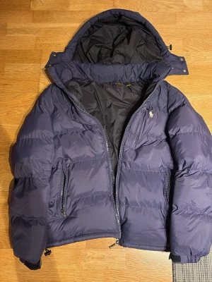 Mörkblå pufferjacka från Polo Ralph Lauren - Snygg mörkblå pufferjacka från Polo Ralph Lauren med huva och dragkedja. Jackan har två framfickor med dragkedja och den klassiska Polo-loggan broderad på bröstet. Perfekt för kalla vinterdagar och riktigt skön att bära.