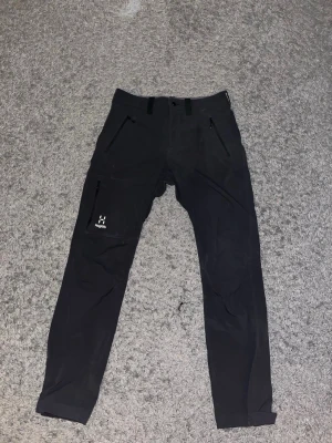 Haglöfs byxor - Svarta byxor från Haglöfs med smal passform, flera dragkedjefickor och logga på benet. Tillverkade i slitstarkt syntetmaterial som funkar perfekt för outdoor och äventyr. Snygg och clean design med bälteshällor och knappstängning. Nypris 1700 kr, nästan oanvända.
