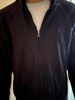 Mörkblå ziptröja från Polo Ralph Lauren - Mörkblå ziptröja från Polo Ralph Lauren med diskret broderad logga på bröstet. Tröjan har ribbad struktur, hög krage och dragkedja framtill. Perfekt för en avslappnad och stilren look.