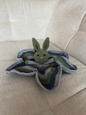 Virkad snuttefilt med kanin - Säljer en handvirkad snuttefilt med kaninmotiv. Filten har stjärnform och är gjord i gröna, blå och grå nyanser. Kaninens huvud och öron är virkat i grönt garn och filten har ett randigt mönster. Perfekt som mysig present till barn.