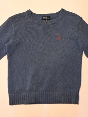 Polo Ralph Lauren blå stickad tröja - Riktigt snygg och stilren blå stickad u ringad tröja från Polo Ralph Lauren med rund halsringning och ribbade muddar. Storlek 152 (12 år). Tröjan har det klassiska röda Polo emblemet broderat på bröstet. Ca 1500 kr nypris. Perfekt för lager på lager, vardagligt eller uppklätt.
