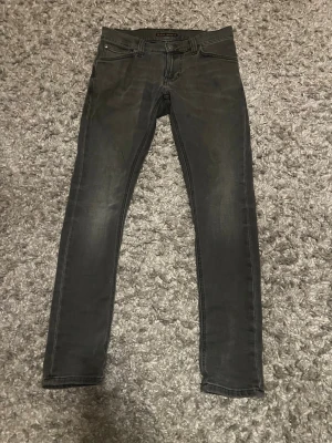 Svarta skinny jeans från Nudie Jeans - Säljer ett par svarta skinny jeans från Nudie Jeans med klassisk femficksdesign och diskreta slitningar. Jeansen har smal passform och stretchigt material för extra komfort. Snygga detaljer på bakfickorna och knappgylf.