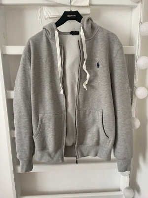 Grå kofta Polo Ralph Lauren Storlek M - Grå huvtröja från Polo Ralph Lauren med dragkedja framtill och luva med vita band. Tröjan har en broderad logotyp på bröstet och muddar vid ärmslut och nederkant. Fickor framtill. Storlek M. Litet hål på luvan och på ryggen som endast syns på väldigt nära håll, annars superfint skick. Knappt använd. Djur, rök och parfymfritt hem.