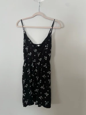 Svart blommig kort klänning - Söt svart kort klänning med tunna axelband och små vita blommor. Klänningen har en lätt v-ringning med knyt framtill och ärmlös design. 