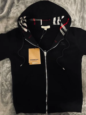 Svart zip hoodie från Burberry - Snygg svart hoodie från Burberry med dragkedja och klassiskt rutigt foder i huvan. Märkets logga är diskret broderad på bröstet. Säljes pga att den inte passade mig, storlek S. Pris kan absolut diskuteras!