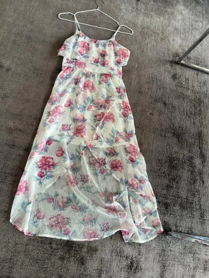 Blommig långklänning med tunna band - Långklänning med tunna axelband och blommigt mönster i rosa och blå toner på vit botten. Klänningen har ett lätt, luftigt tyg och ett lager över bysten för extra detalj. Perfekt för varma dagar och somriga tillfällen.