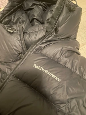 Svart dunjacka Peak Performance - Säljer en svart Peak Performance jacka med logga på bröstet och ärmen. Jackan har dragkedja, hög krage och justerbara snören vid huvan. Klassisk puffer-modell i slitstarkt material.