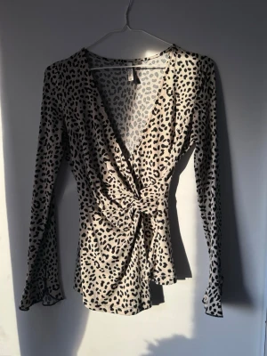 Leopardmönstrad omlottopp med knyt - Snygg leopardmönstrad omlottopp i beige och svart med lång ärm och knyt i midjan. Djup v-ringning och figurnära passform. Perfekt för dig som vill sticka ut lite extra. Materialet känns mjukt och stretchigt, troligen polyester.