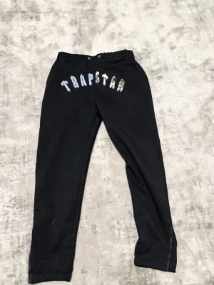 Svarta Trapstar mjukisbyxor med dragkedja - Säljer ett par svarta mjukisbyxor från Trapstar med logga i vitt och blått framtill. Byxorna har resår i midjan med snörning och dragkedjor vid bensluten för extra stil. Perfekta för en chill och trendig look. 