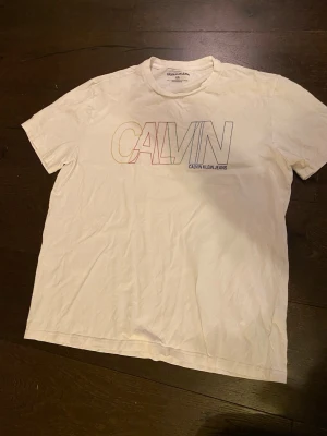 Vit Calvin Klein Jeans t-shirt - Vit t-shirt från Calvin Klein Jeans med stort, färgglatt CALVIN-tryck på bröstet i regnbågsfärger. Klassisk rund halsringning och korta ärmar. Tillverkad i mjuk bomull för en skön och avslappnad känsla.