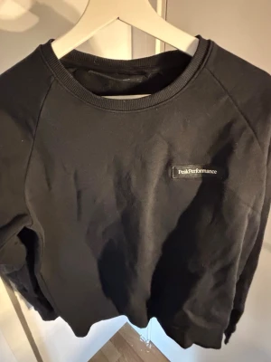 Svart sweatshirt Peak Performance M - Klassisk svart sweatshirt från Peak Performance med rund halsringning och diskret logga på bröstet. Tröjan har ribbade muddar vid ärmslut och nederkant. Perfekt för en avslappnad och stilren look.