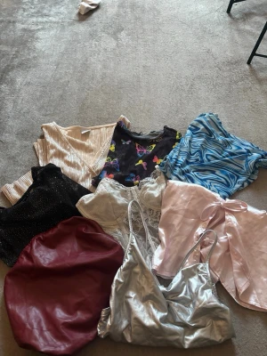 Mixade toppar i olika färger och material - Säljer ett gäng snygga toppar i olika stilar: silvrig camitopp i syntet, röd topp i skinnimitation, svart glittrig topp, beige ribbad topp, blå mönstrad topp, vit spetslinnetopp, rosa satinlinnetopp och en svart topp med färgglada fjärilar. Perfekt för att variera din look!