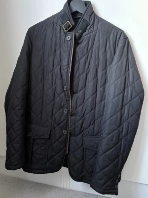 Barbour jacka - Riktigt stilren svart quiltad jacka från Barbour i storlek S med hög krage och spänne i halsen. Jackan har knapp- och dragkedjestängning, två framfickor med lock och en innerficka. Passar utmärkt nu till våren! Inga defekter! Bara att kontakta vid funderingar!🌟