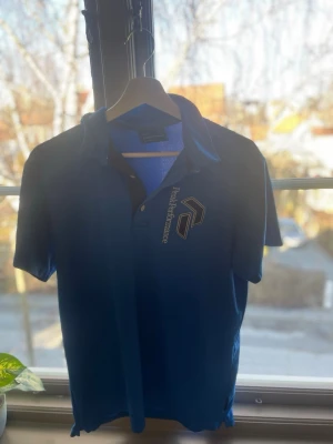 Blå Peak Performance piké - Snygg blå pikétröja från Peak Performance med klassisk krage och korta ärmar. Tröjan har logga tryckt på bröstet och är gjord i ett mjukt och bekvämt material, perfekt för en avslappnad stil. Storleken är L men det är liten i storleken så passar perfekt till dem som har storlek m och s beroende på hur du vill ha passformen!