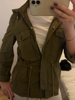 Olivgrön parkajacka med huva - Snygg olivgrön parkajacka med huva och dragsko i midjan. Jackan har flera fickor framtill, dragkedja och knappar samt långa ärmar. Perfekt för dig som vill ha en stilren och praktisk jacka till vardags. Knappen på ena ärmen är av men det går såklart o vika upp ändå.