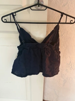 Svart broderad linnetopp med spets - Söt mörkblå linnetopp med tunna axelband, v-ringning och broderade detaljer. Toppen har spetskant, knyt framtill och är croppad i modellen. Storlek Xs och från Subdued.