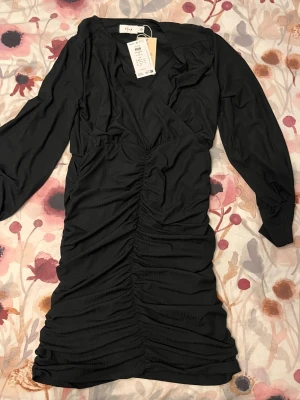Svart klänning från Vila - Svart klänning från Vila med långa ärmar och snygg rynkning. Klänningen har v-ringad hals och är tillverkad i ett mjukt, lätt material som ger en smickrande passform. Perfekt för dig som gillar stilrena och trendiga plagg.