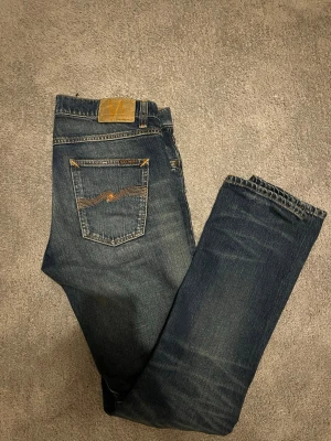 Nudie jeans  - Säljer ett par klassiska blå jeans från Nudie Jeans med snygga slitningar och kontrastsömmar. Modellen har normal passform och fem fickor, samt Nudie Jeans ikoniska broderi på bakfickan. Perfekta till en avslappnad stil. Feta slitningar fint skick storlek 31/34