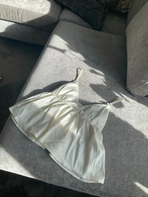 Vit linnetopp med tunna axelband - Säljer en söt vit linnetopp med tunna axelband decorerade med rosetter. Toppen har en lätt utsvängd passform och är gjord i ett mjukt, glansigt material som känns lyxigt. Perfekt för varma dagar eller att styla lager på lager.