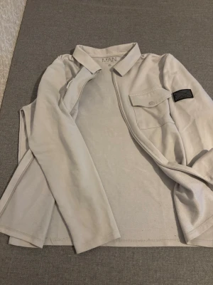 Grå overshirt  - Stilren grå overshirt  med dragkedja framtill, klassisk krage och en bröstficka med knapp. På vänster ärm sitter en svart patch med logga. Overshirten är tillverkad i ett mjukt bomullsliknande material och har raka ärmar.