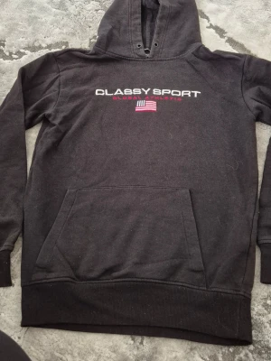 Svart hoodie Classy Sport - Svart hoodie från Classy Sport med trycket 'Classy Sport Global Athletic' och en amerikansk flagga på bröstet. Klassisk känguruficka framtill och huva med dragsko. Perfekt för en avslappnad och sportig stil.