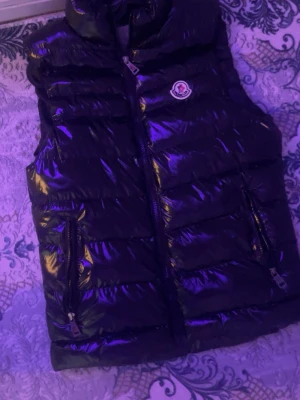 Svart glansig dunväst från Moncler - Snygg svart dunväst från Moncler med glansig finish och hög krage. Västen har två dragkedjefickor framtill och klassisk Moncler-logga på bröstet. Perfekt för lager-på-lager och streetstyle.