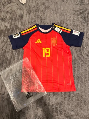 Spaniens matchtröja Lamine Yamal 19 - Röd och marinblå Spanien fotbollströja från Adidas med gula detaljer, nummer 19 och Lamine Yamal på ryggen. Tröjan har korta ärmar, tunna vertikala ränder och officiellt landslagsemblem på bröstet. Perfekt för fotbollsfans som vill stötta sitt lag.