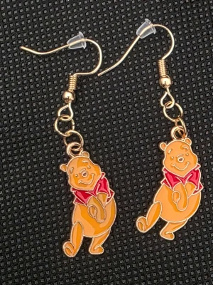 Örhängen med Nalle Puh-motiv - Säljer ett par unika örhängen med motiv av Nalle Puh i gul och röd emalj. Hängena är i metall med krokfäste och plastpluppar. Perfekta för dig som älskar söta och lekfulla accessoarer med Disney-tema.
