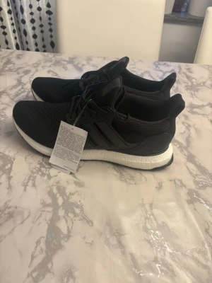 Svarta Addidas ultra boost  med vit sula - Stilrena svarta sneakers med vit sula och stickad ovandel. Skorna har snörning och en sportig design med bekväm dämpning. Perfekta för dig som gillar en clean look och vill ha något som funkar till det mesta. Materialet är mesh och syntet.