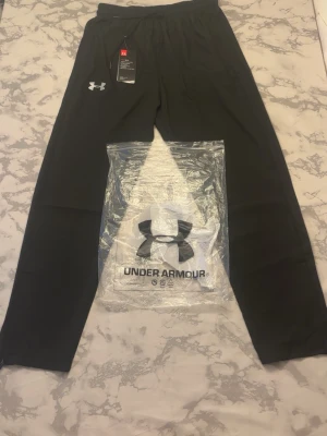 Svarta mjukisbyxor från Under Armour - Snygga svarta mjukisbyxor från Under Armour med resår i midjan och dragsko. Byxorna har en diskret logga på vänster lår och är tillverkade i ett mjukt och stretchigt material som passar perfekt för chill eller träning.