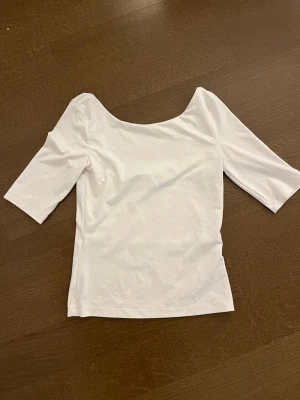 Vit topp med halvlånga ärmar - En stilren vit topp med halvlånga ärmar och bred, rundad urringning både fram och bak. Tillverkad i mjukt bomullsmaterial som känns skönt mot huden. Perfekt att matcha med jeans eller kjol för en clean look.