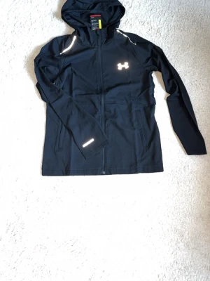 Svart vindjacka från Under Armour - Snygg svart vindjacka från Under Armour med huva och hel dragkedja framtill. Jackan har reflexdetaljer på axlar och ärm, samt två sidofickor. Perfekt för blåsiga dagar och sportiga aktiviteter.