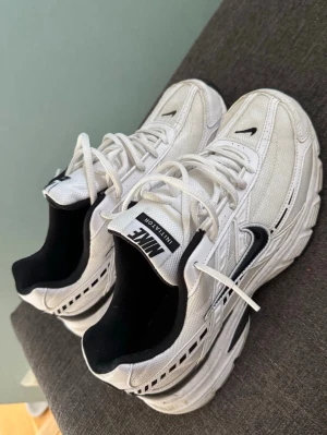 Vita Nike sneakers  - Snygga vita sneakers från Nike med svarta detaljer och klassisk swoosh-logga på sidan. Skorna har snörning och en chunky sula som ger en sportig vibe. Ovandelen är i mesh och syntet för bra andningsförmåga och komfort.