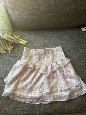 Rosa blommig volangkjol från Gina Tricot - Supersöt kort kjol med blommigt mönster i rosa och vitt. Kjolen har bred smock i midjan och dubbla volanger som ger en luftig och lekfull känsla. Perfekt för varma dagar och enkel att matcha med en enkel topp.