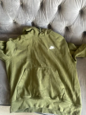 Olivgrön hoodie från Nike - Snygg olivgrön hoodie från Nike med klassisk logga broderad i vitt på bröstet. Tröjan har en stor känguruficka framtill och huva utan snören. Perfekt för en avslappnad streetwear-look.