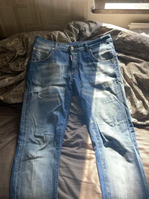 Blå slitna jeans från Dsquared2 - Säljer ett par blå jeans från Dsquared2 med slitningar och distressed detaljer på benen. Klassisk femficksmodell med knappgylf och läderpatch bak i midjan. Perfekta för en avslappnad och trendig look.