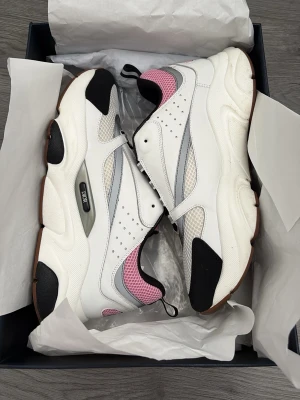 Chunky vita sneakers med rosa detaljer - Säljer ett par chunky sneakers i vitt med svarta och rosa detaljer samt grå inslag. Skorna har en tjock sula, meshpaneler och perforerade partier för extra stil. Perfekta för dig som gillar streetwear och vill sticka ut lite extra.