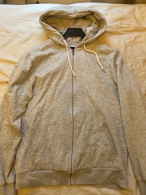 Ljusgrå zip hoodie med huva - En enkel och stilren ljusgrå hoodie med dragkedja framtill och två fickor. Hoodien har justerbar huva med vita snören och ribbade muddar vid ärmslut och nederkant. Perfekt för en avslappnad look.