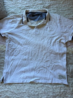 Vit pikétröja med broderad logga - Klassisk vit pikétröja med krage och knappar framtill. Tröjan har en diskret broderad logga på bröstet och mörka detaljer på kragen och ärmsluten. Tillverkad i bomull för en bekväm känsla. Perfekt till jeans eller shorts.