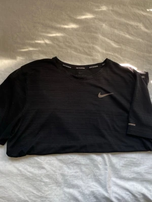 Nike Dri-Fit  - Säljer en svart Nike Dri-Fit löpartröja med rund hals och korta ärmar. Tröjan har Nike-logga på bröstet och är tillverkad i ett lätt, snabbtorkande funktionsmaterial som passar perfekt till träning och löpning.