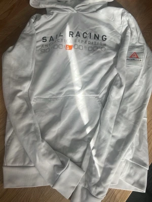 Vit hoodie Sail Racing Antarctica - Vit hoodie från Sail Racing med tryck 'Antarctica Expedition' på bröstet och patch på ärmen. Klassisk huva med snörning och känguruficka framtill. Perfekt för dig som gillar stilren och sportig look.