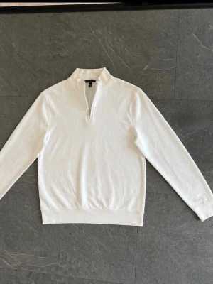 Vit halvzip tröja med hög krage - Snygg vit långärmad tröja med halvzip och hög krage. Tröjan har ribbade muddar vid ärmslut och nederkant. Perfekt för en clean och stilren look. Materialet ser ut att vara mjukt och bekvämt, troligen bomull eller bomullsblandning.