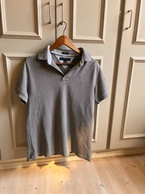 Grå pikétröja från Tommy Hilfiger - Klassisk grå pikétröja från Tommy Hilfiger med liten logga på bröstet. Tröjan har korta ärmar, krage och knappar vid halsen. Perfekt för en stilren och avslappnad look.
