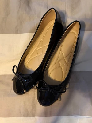 Svarta lackade ballerinaskor med rosett - Stilrena svarta ballerinaskor i glansigt lackmaterial med rund tå och dekorativ rosett framtill. Skorna har en mjuk, vadderad innersula i beige och låg klack. Perfekta för dig som gillar klassisk och enkel stil.