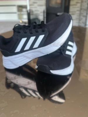 Svarta Adidas sneakers med vita ränder - Snygga svarta sneakers från Adidas med klassiska vita ränder på sidan och tjock vit sula. Ovandelen är i mesh-tyg för bra andningsförmåga och komfort. Perfekta för dig som vill ha en sportig och stilren look.