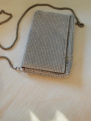 Silvrig kuvertväska med kedja - Snygg kuvertväska täckt av glittrande silvriga stenar och med en matchande silverfärgad kedjerem. Väskan har en rektangulär form och stängs med lock. Perfekt för att addera lite extra bling till din outfit.