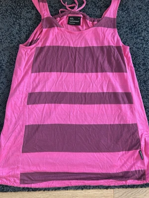 Rosa och mörkrosa randig linnetopp - Säljer en snygg randig linnetopp i rosa och mörkrosa toner. Toppen har breda axelband och ärmlös design, perfekt för varma dagar. Tillverkad i mjuk bomull som känns skön mot huden. Passar dig som gillar färg och vill sticka ut lite extra.