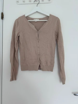 Beige tunn kofta med knappar - En stilren beige kofta med v-ringning och knäppning framtill. Långärmad modell med ribbade muddar och nederkant. Perfekt att slänga över en t-shirt eller linne för en enkel och clean look.