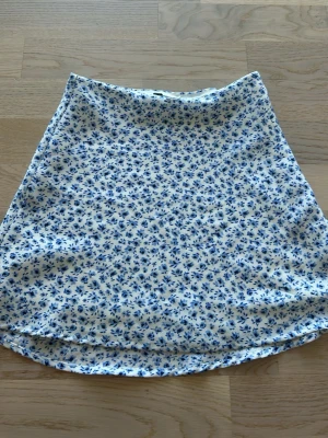Vit-blå blommig kortkjol - Söt kortkjol med blått blommönster på vit botten. Kjolen har en diskret dragkedja i sidan för enkel på- och avtagning. Perfekt för dig som gillar en feminin och fräsch stil. Materialet känns mjukt och lätt, perfekt för varmare dagar.